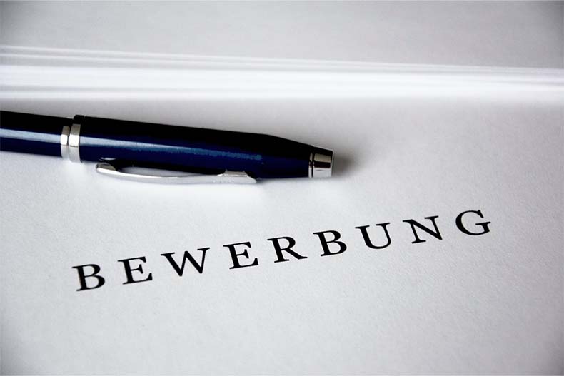 Ein blauer Kugelschreiber liegt auf einem weißen Blatt Papier, auf dem mit schwarzen Buchstaben das Wort "Bewerbung" steht.
