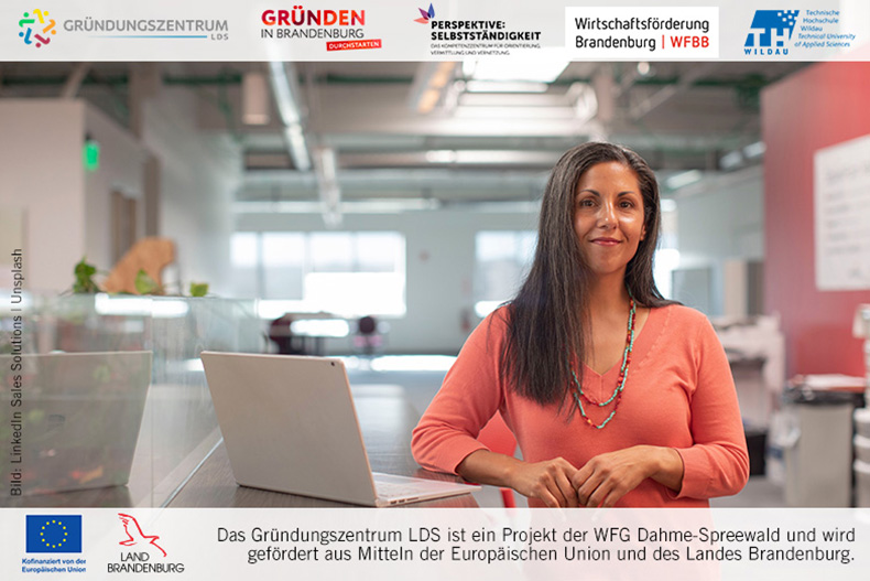 Eine Frau mit einem orangefarbenen Shirt steht vor einem Laptop.