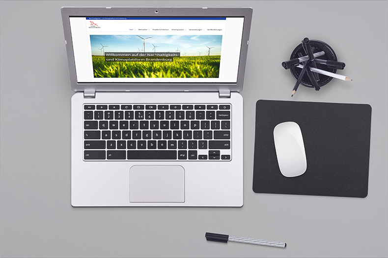 Ein Laptop, der die Homepage des Nachhaltigkeits- und Klimaplattform zeigt. Um den Laptop herum befinden sich ein Stift, ein Stifteköcher sowie ein Mauspad samt Maus. 