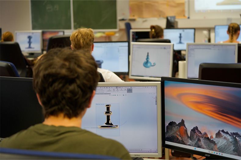 Ein junger Mann sitzt zusammen mit weiteren Studierenden im Hörsaal einre Hochschule. Vor ihm steht ein aufgeklapptes Notebook.