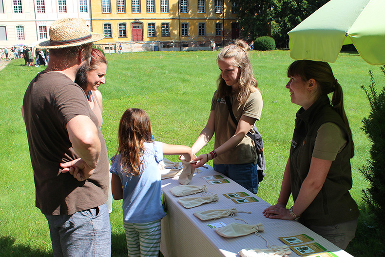 Ein Mann, eine Frau und ein Mädchen stehen an einer Station der Naturwelt-Olympiade beim Sommerfest der Lieberoser Heide 2025. Das Mädchen fasst in einen Jute-Beutel, um zu erraten, welche Frucht und welches Laubblatt zu welchem Baum gehören. 